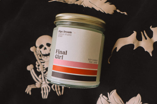 A candle labeled Final Girl staged on top of a black and white ghost sweatshirt.
