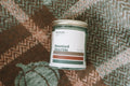A glass jar candle labeled 'Haunted Hayride' by Pipe Dream Candle, placed on a flannel green and orange blanket with pumpkins. 