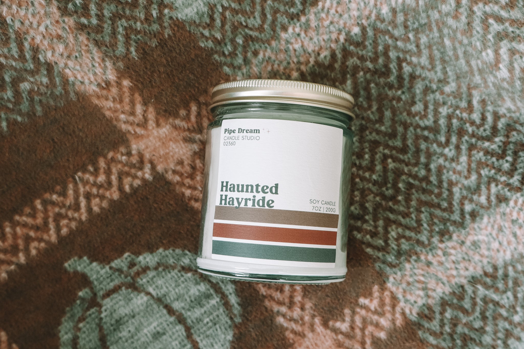 A glass jar candle labeled 'Haunted Hayride' by Pipe Dream Candle, placed on a flannel green and orange blanket with pumpkins. 