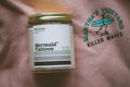Candle labeled 'Mermaid Tattoos' on a surface with a shirt featuring 'Martha's Vineyard Killer Waves' design.