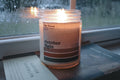Candle labeled 'October Rain' on a surface with a rain covered window in the background