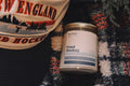 Candle labeled 'Pond Hockey' on a textured surface with a New England hockey hat in the background.