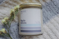 A jar candle with a label that reads 'Rain Boots', accompanied by a few withered yellow flowers, all placed on a grey textured surface.